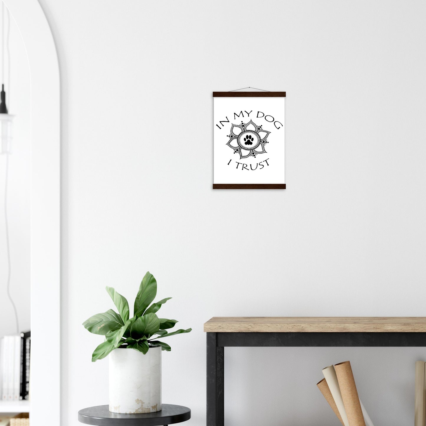 Poster with 'In my dog I trust' text around a paw print mandala design with a Light colored hanging poster frame with a top and a bottom only on a white wall above a plant and desk.